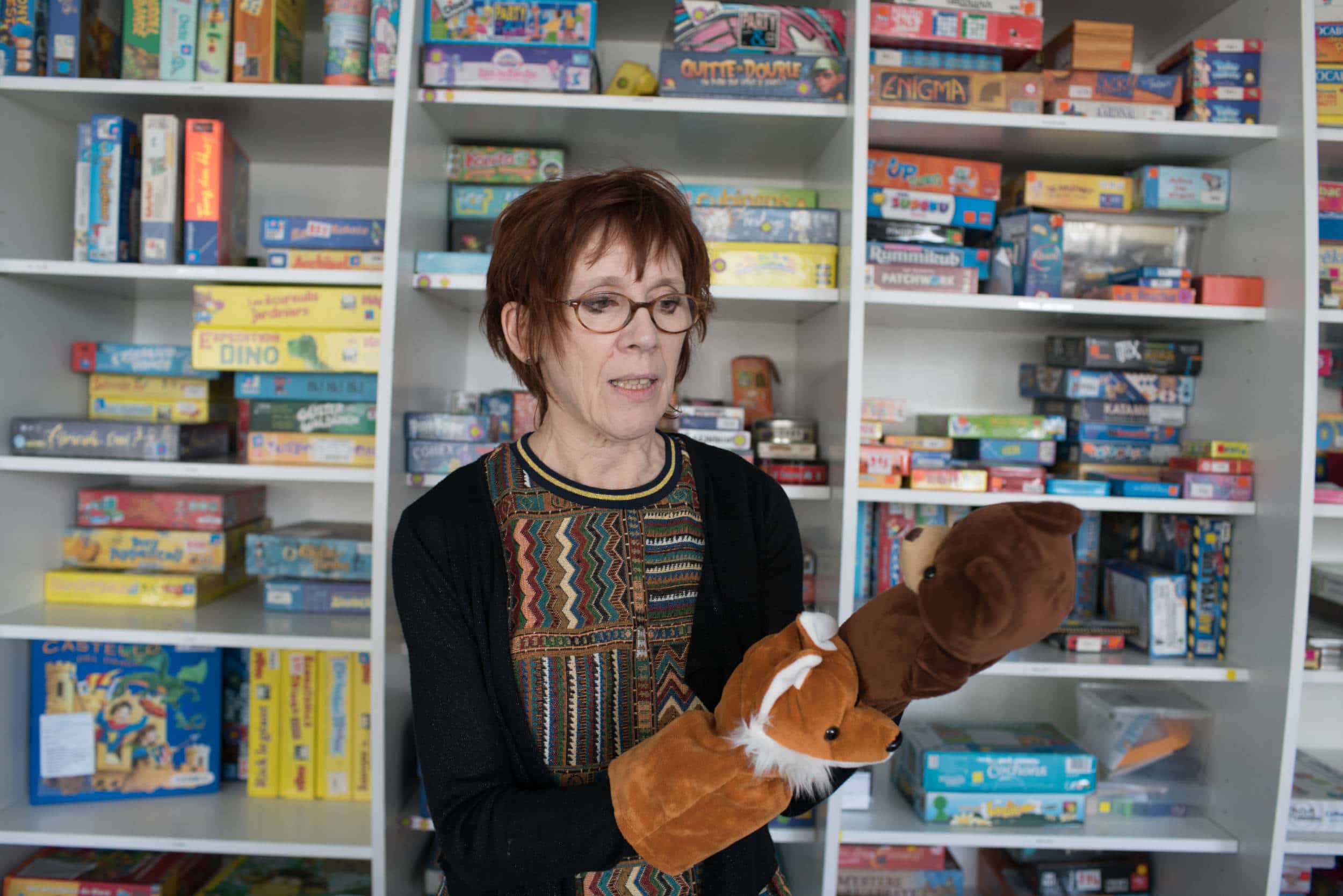 Devant une ludothèque remplie de jeux de société, Isabelle montre deux marionnettes d'ours et de renard qu'elle a enfilées sur ses mains
