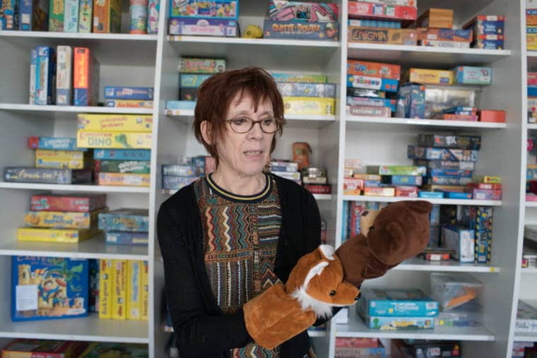 Devant une ludothèque remplie de jeux de société, Isabelle montre deux marionnettes d'ours et de renard qu'elle a enfilées sur ses mains