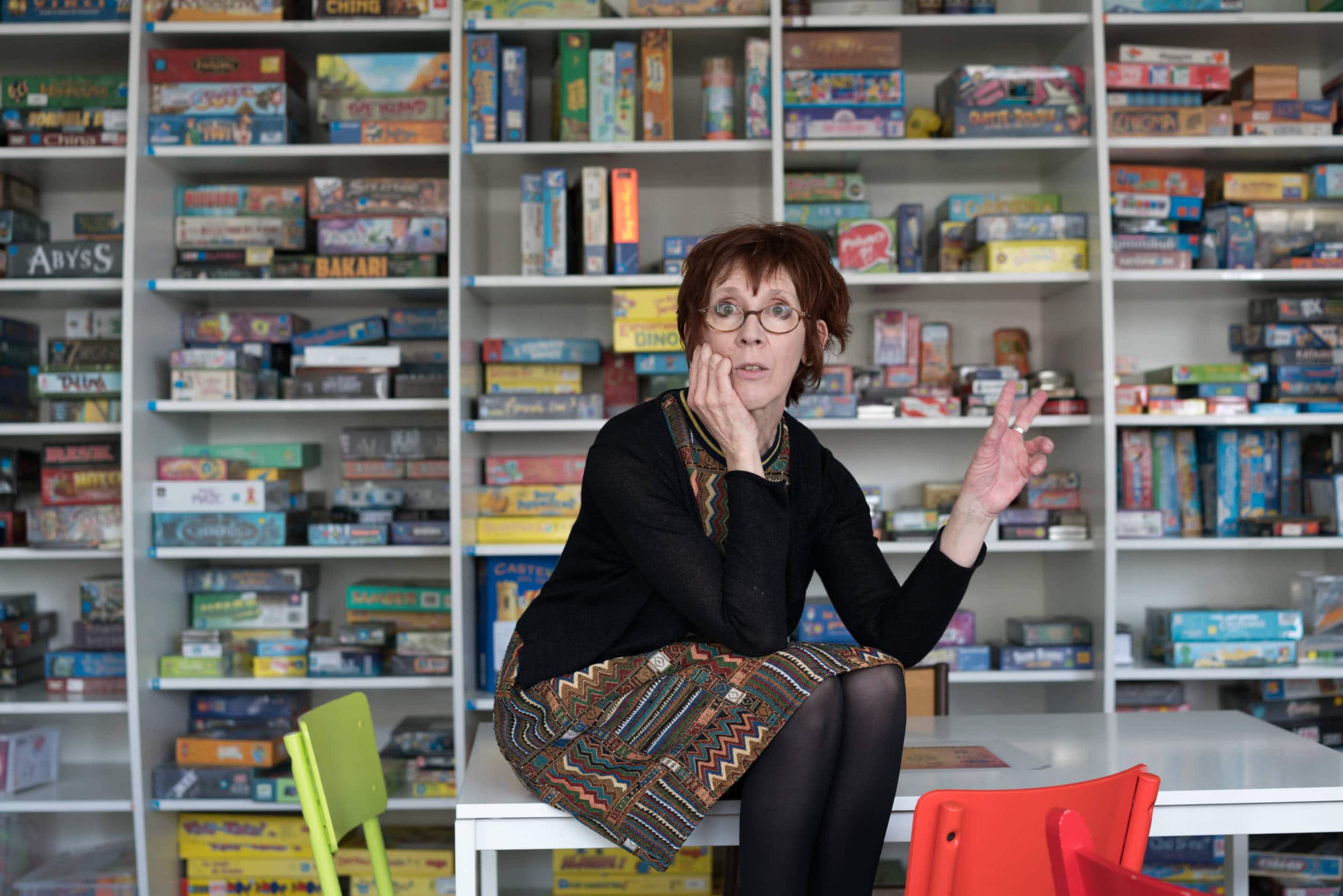 Devant la ludothèque, Isabelle est assise sur un bureau et s'adresse à un auditoire invisible, de face