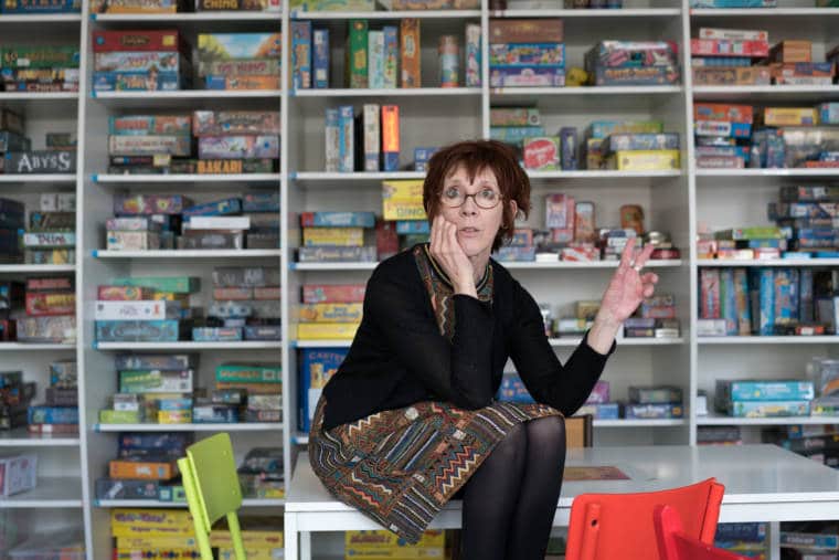 Devant la ludothèque, Isabelle est assise sur un bureau et s'adresse à un auditoire invisible, de face