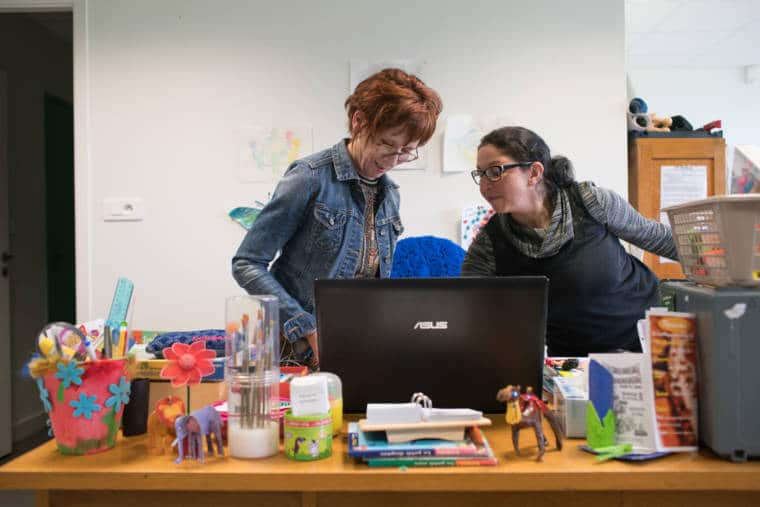 Derrière un ordinateur portable, sur un bureau orné de décorations dans le thème de l'enfance, Isabelle et une collègue, souriantes, travaillent
