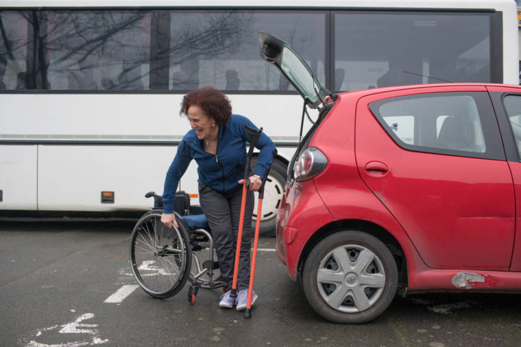 Devant le coffre de sa voiture rouge, Hadda, souriante, s'appuie sur ses béquilles pour se hisser dans son fauteuil.