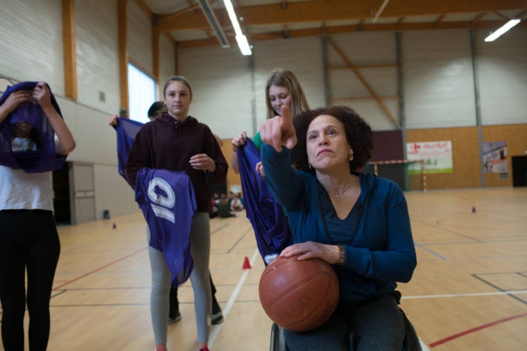 De face, un ballon de basket à la main, Hadda désigne un objet que l'on ne voit pas à ses élèves.