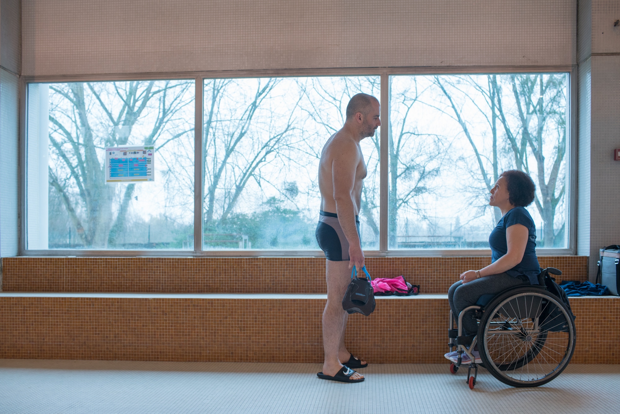 Hadda fait face à un nageur adulte devant une grande vitre donnant sur l'extérieur de la piscine.