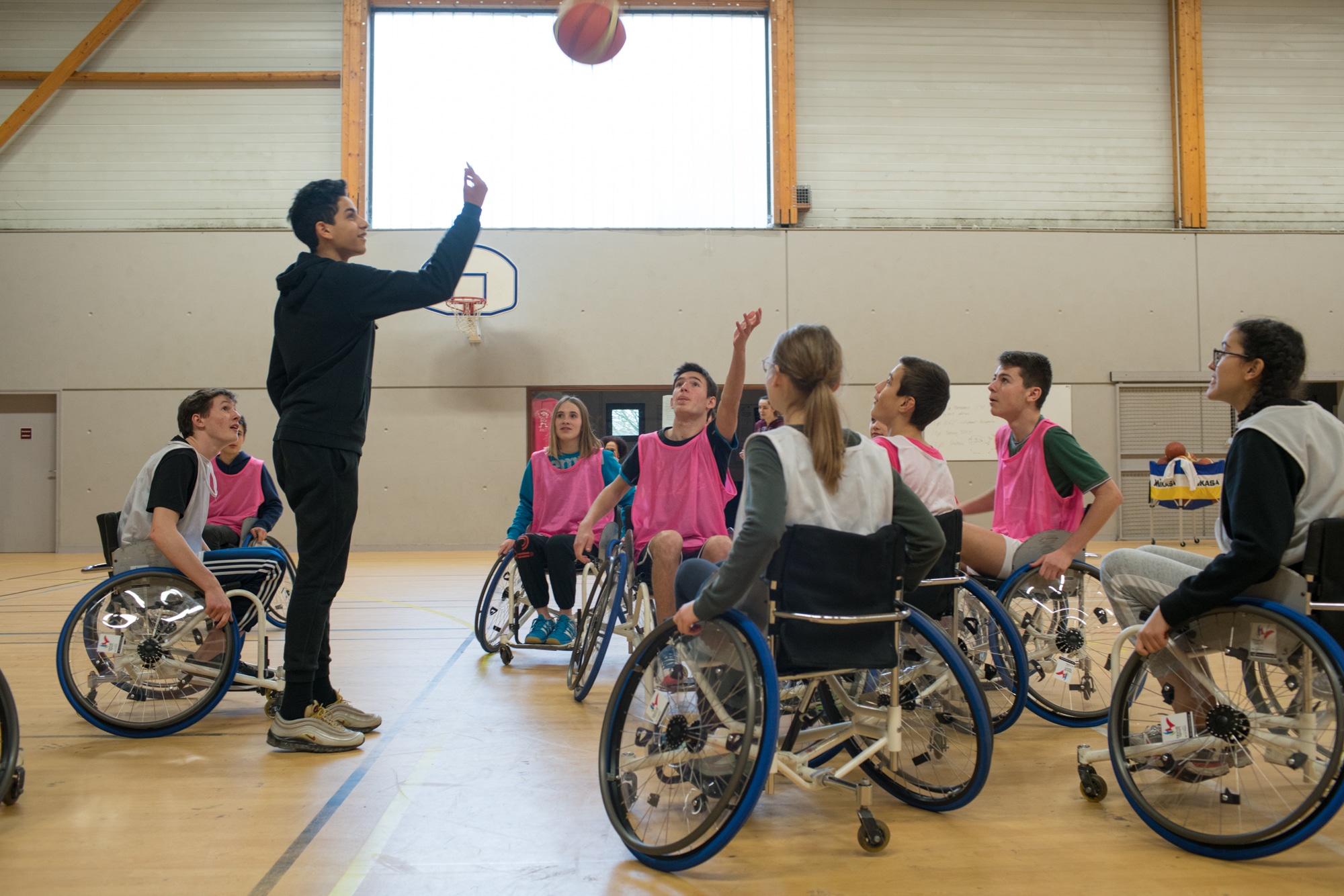Un élève, debout, lance un ballon pour engager un entre-deux à des élèves en fauteuil.