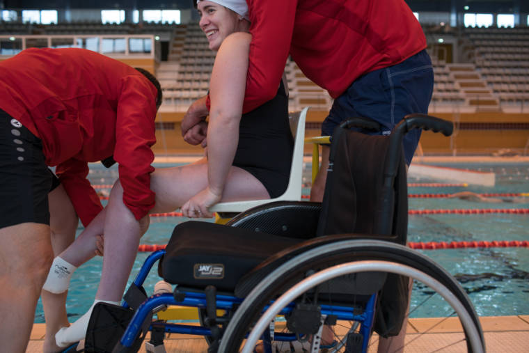 Une élève d'Haddam, équipée d'un maillot et d'un bonnet, est aidée par deux personnes pour s'extirper de son fauteuil roulant.