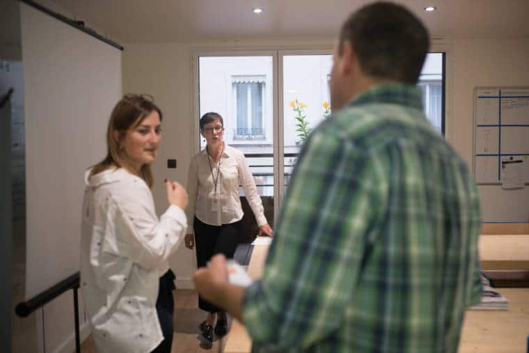 Mathilde, dos à la fenêtre, positionnée près d'un écran de projection, s'adresse à deux personnes.